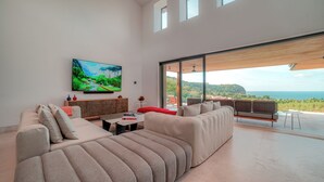 A modern living room with a large sofa, a television, and a view of the ocean through large windows.