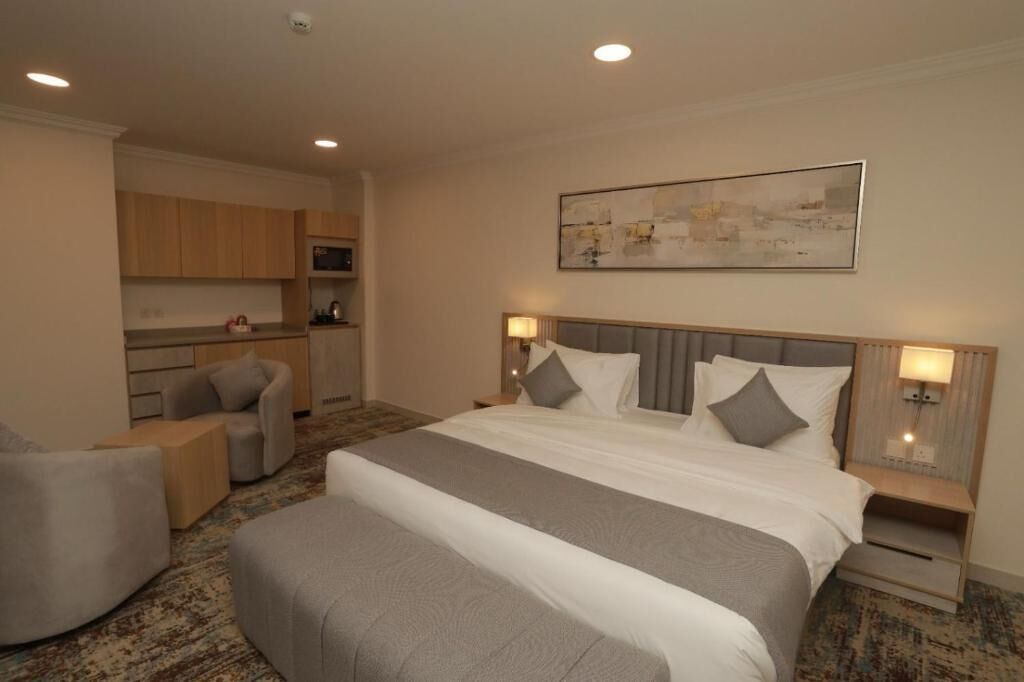 A hotel room with a large bed, bedside tables, a seating area with chairs, a small kitchenette, and a painting on the wall.