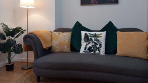 A living room with a grey sofa, a potted plant, a floor lamp, and a framed artwork on the wall.