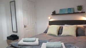 A neatly made bed with multiple pillows and folded towels, a bedside table with a lamp, a wall-mounted shelf with decorative items, and a door.