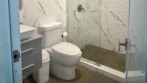 Un baño con ducha envuelta en vidrio, inodoro blanco y tocador con lavamanos y cajones.