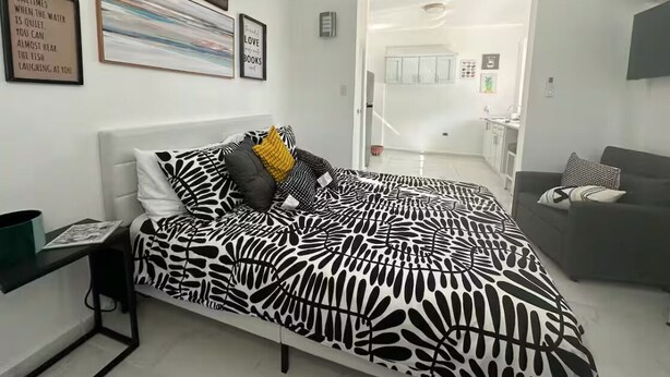 Un dormitorio moderno con cama, mesita de noche, sofá y una cocina visible a través de una puerta abierta.