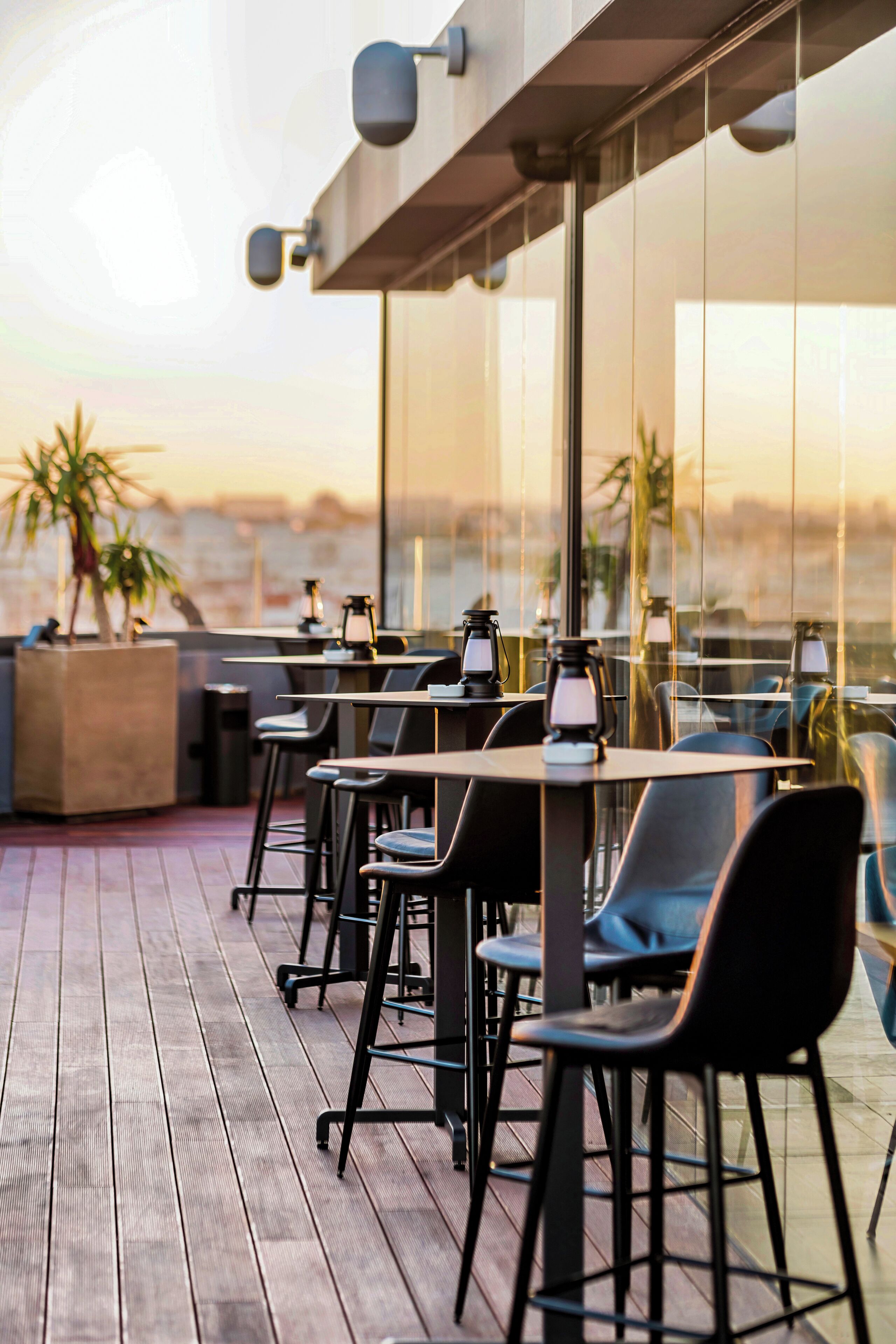 A rooftop terrace with tables and chairs, large windows, and potted plants.