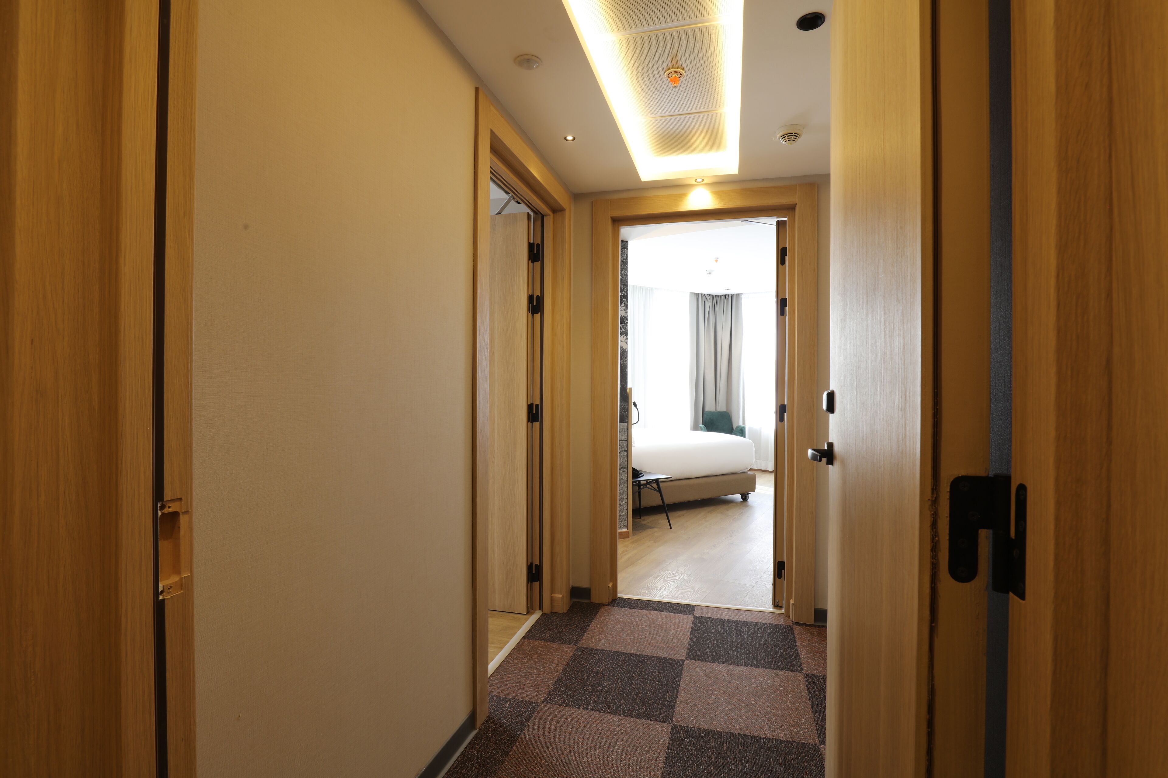 A hotel hallway with wooden doors, a checkered floor, and a bed in the distance.