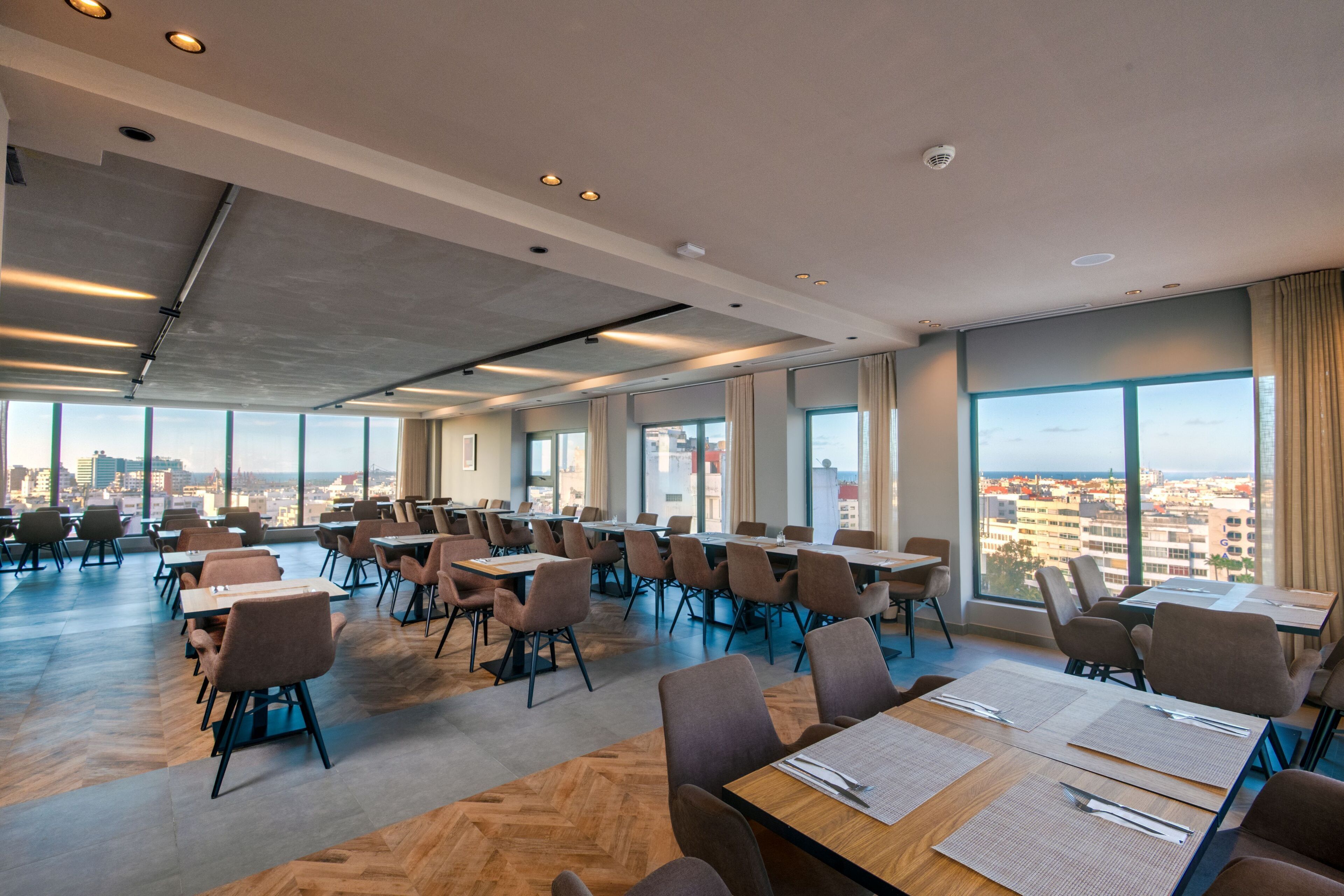 A spacious conference room with large tables and chairs, large windows offering a city view, and a modern ceiling design.