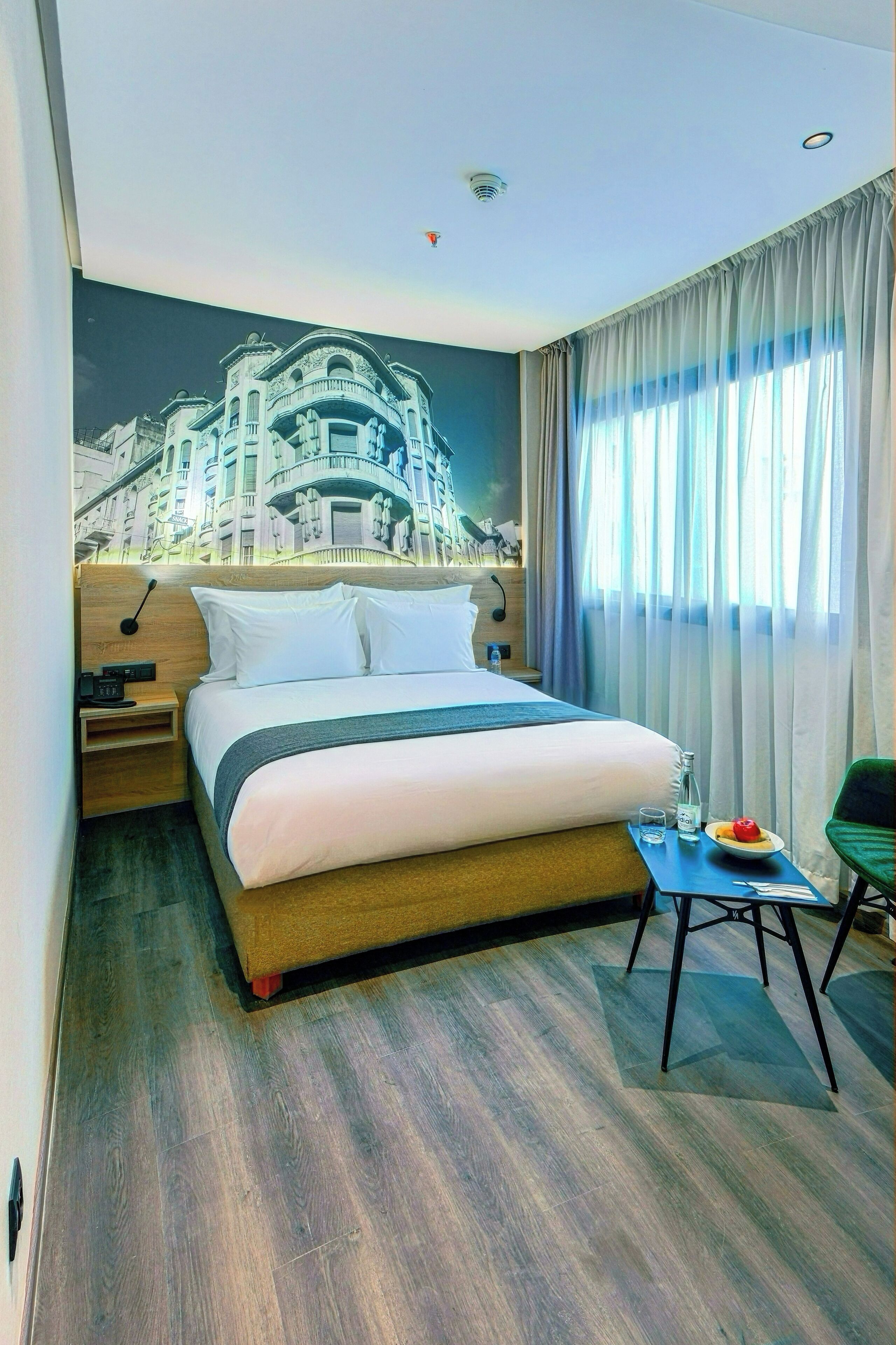 Habitación de hotel con una cama grande, una mesita de noche, una pequeña mesa con un plato y un vaso, y una silla.