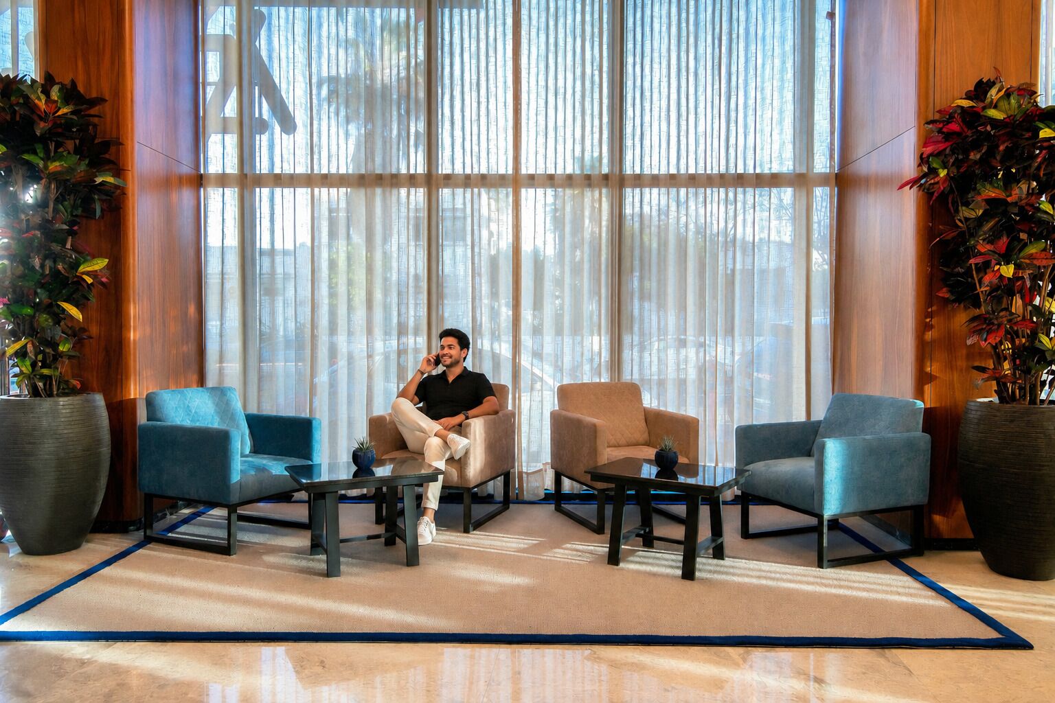 A man sitting on a beige armchair in a lobby with large windows, two blue armchairs, and potted plants.