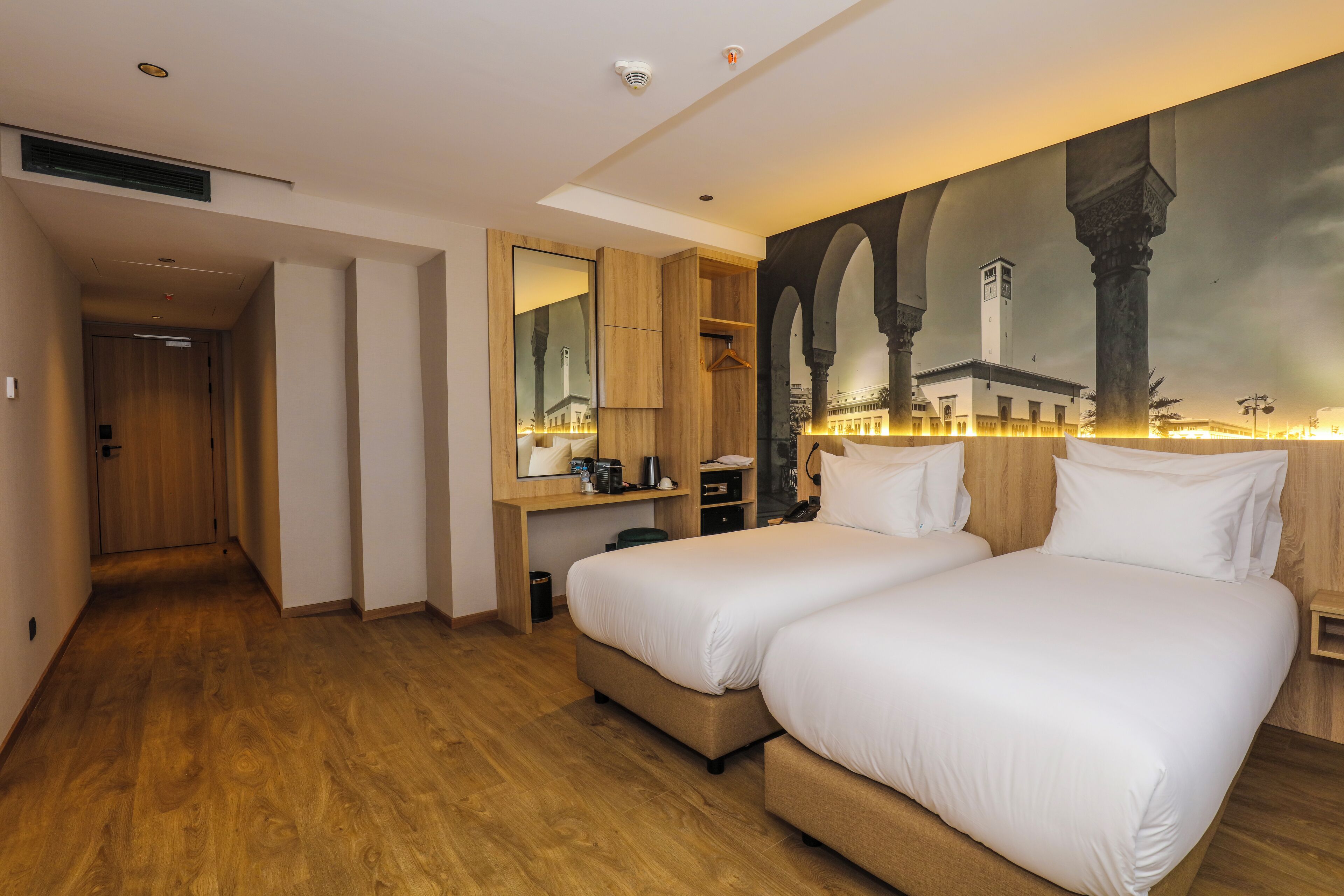 Habitación de hotel con dos camas, piso de madera, un espejo grande, televisor montado en la pared y una pintura de una mezquita.
