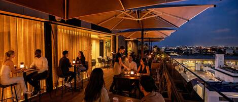 An outdoor dining area with tables and chairs, umbrellas, and a view of the cityscape.