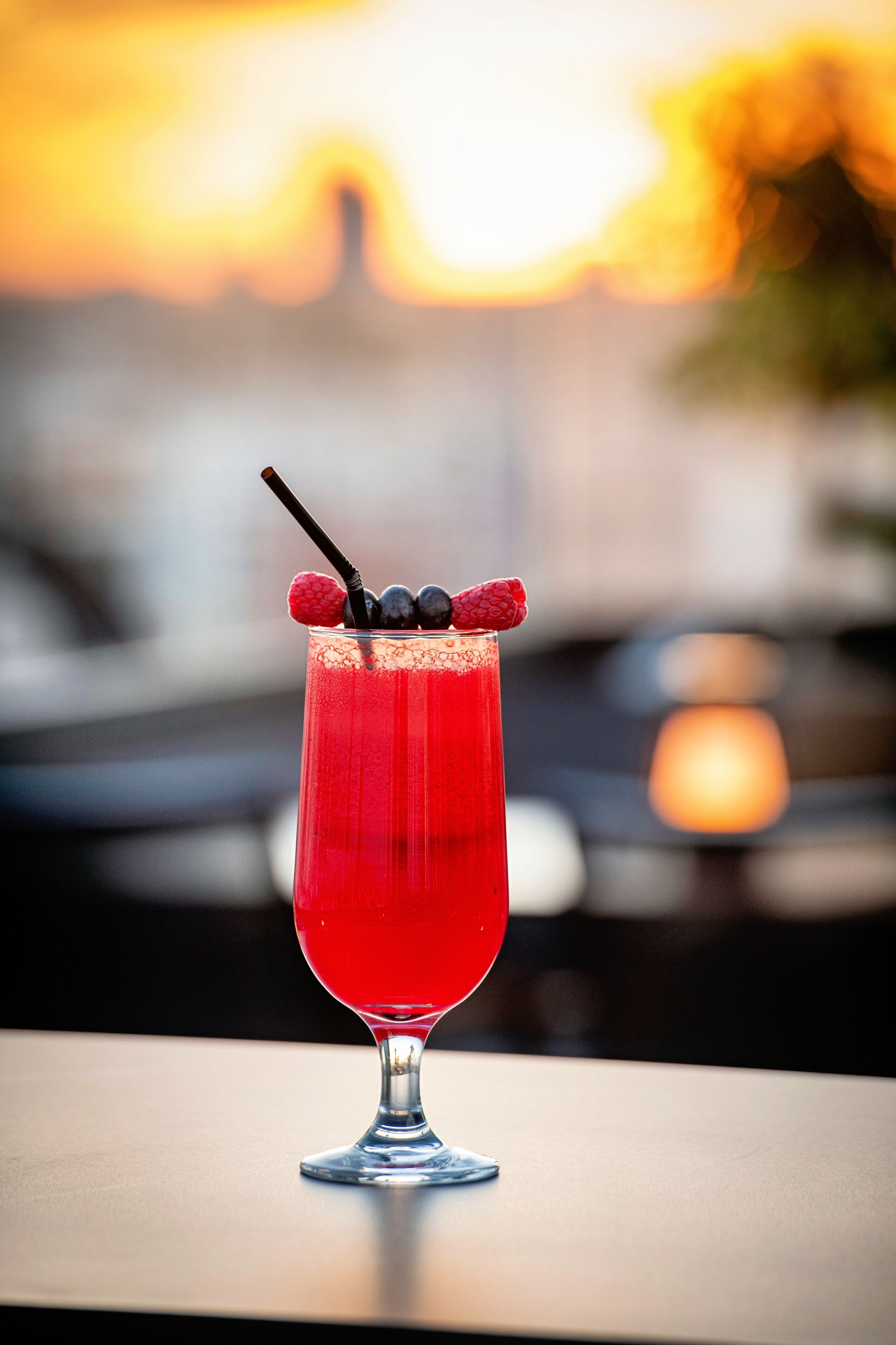 A tall glass with a red beverage, garnished with raspberries and a straw, on a table with a blurred cityscape background.