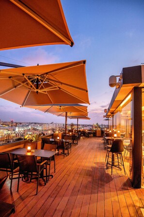 Eine Terrasse im Freien mit Holzboden, Tischen, Stühlen und großen Sonnenschirmen, die Schatten spenden.