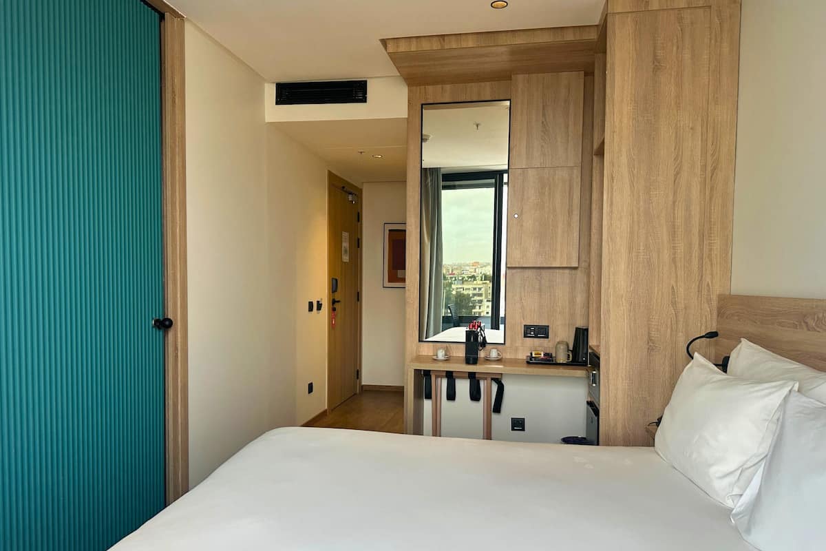 A hotel room with a bed, a mirror, a desk, and a view of the city.