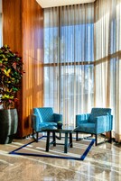 Un vestíbulo moderno con dos sillones azules, una mesita de centro, una planta grande en maceta y un ventanal con cortinas traslúcidas.