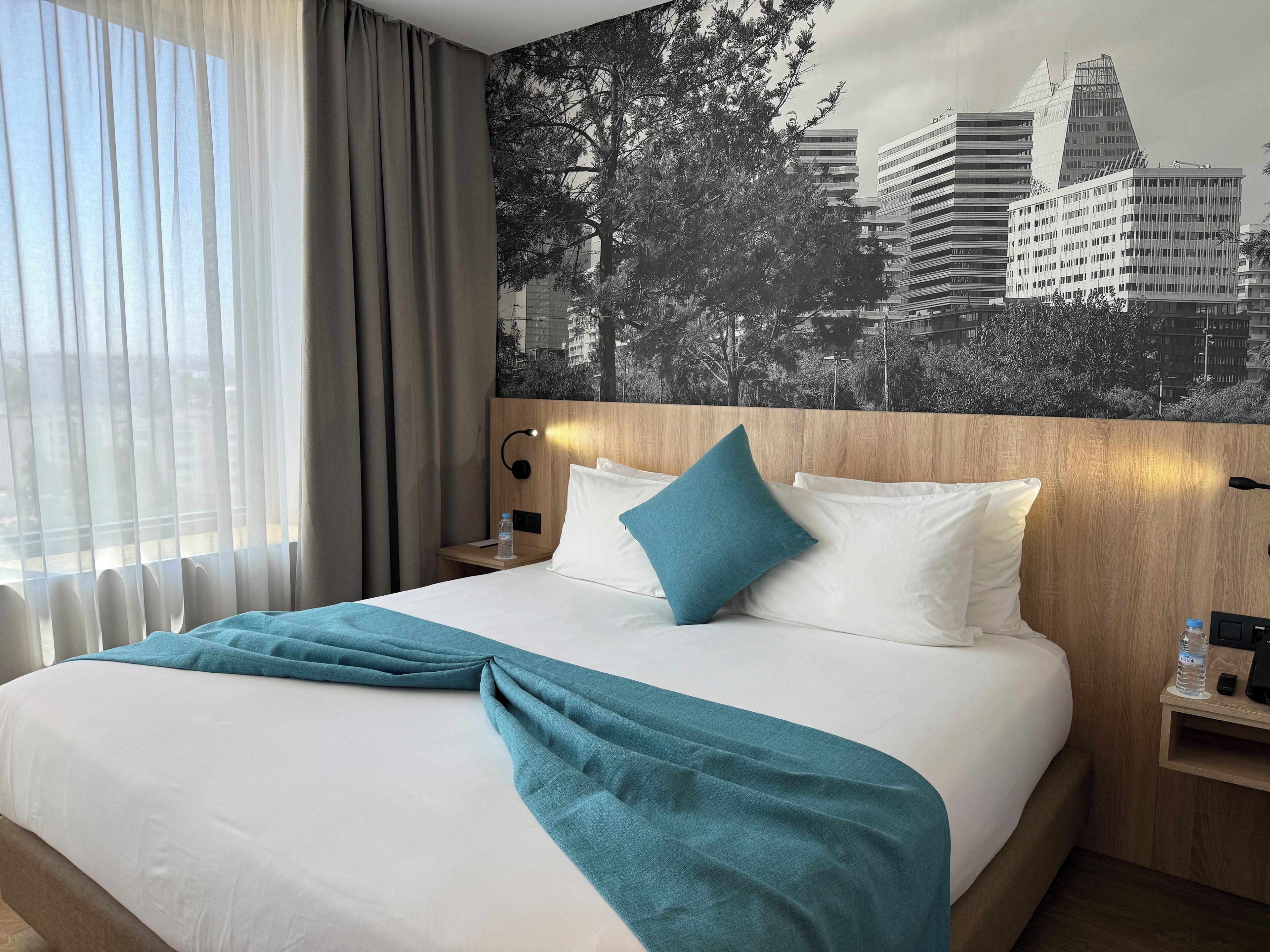 Habitación de hotel con cama, mesita de noche, lámpara de pared, botella de agua y un mural con vista a la ciudad.