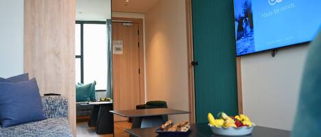 A modern hotel room with a sofa, a dining table with fruit, and a TV displaying 'DW Made for minds.'