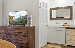 Quarto com cômoda de madeira, televisão em cima, porta branca, banheiro com pia e espelho, e um tapete pequeno no chão.