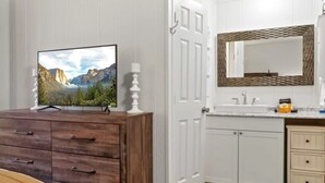 Une salle de bain avec un meuble-lavabo en bois, un grand miroir, un lavabo et un téléviseur diffusant un paysage de montagne.