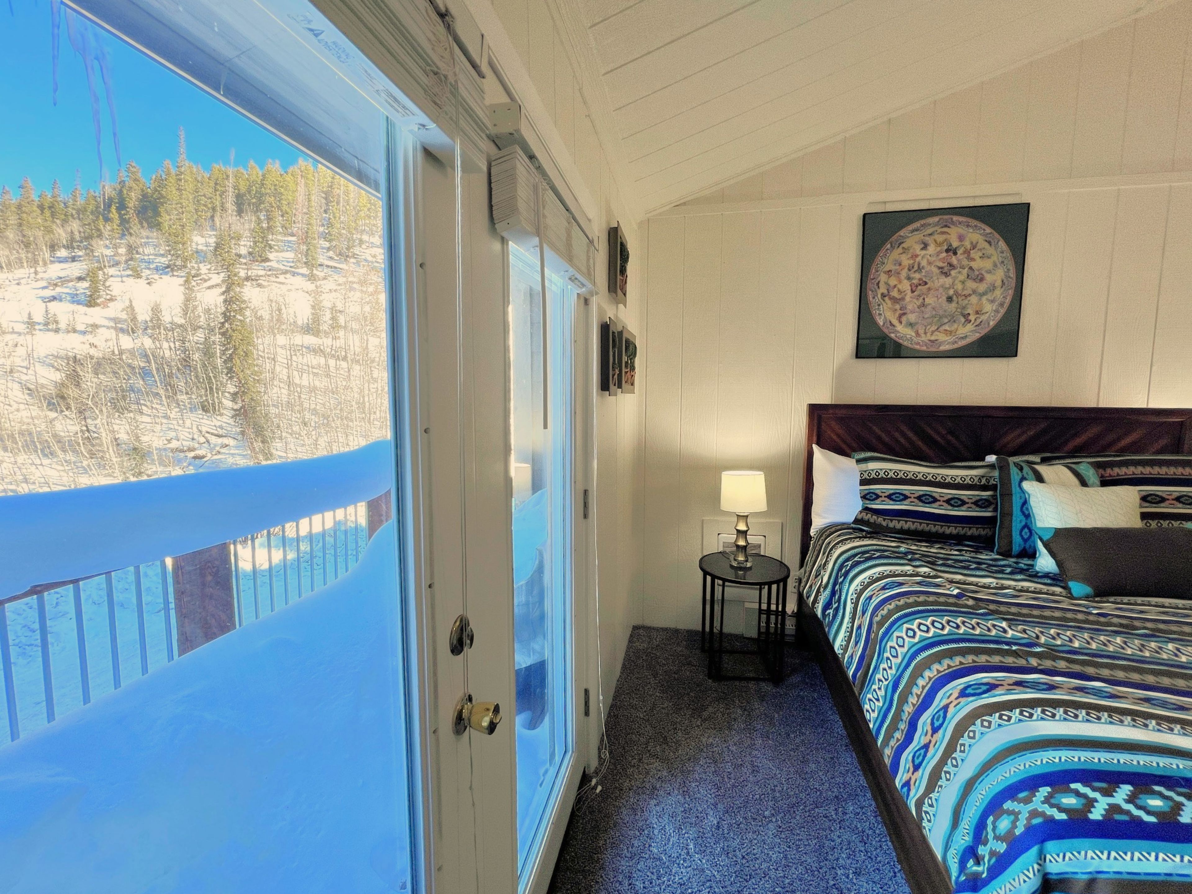 Una habitación con cama, una mesita de noche con lámpara, una ventana con vistas a árboles y nieve, y una pared con un cuadro enmarcado.