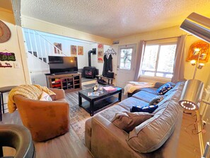 A living room with a sofa, armchair, coffee table, TV, fireplace, and a staircase leading upstairs.