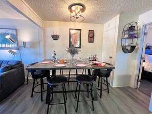 A dining area with a table set for four, surrounded by black chairs, a vase with flowers, and a painting on the wall.