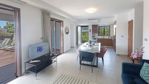 A modern living room with a TV, dining table, and chairs. There is a sofa and a kitchen in the background.