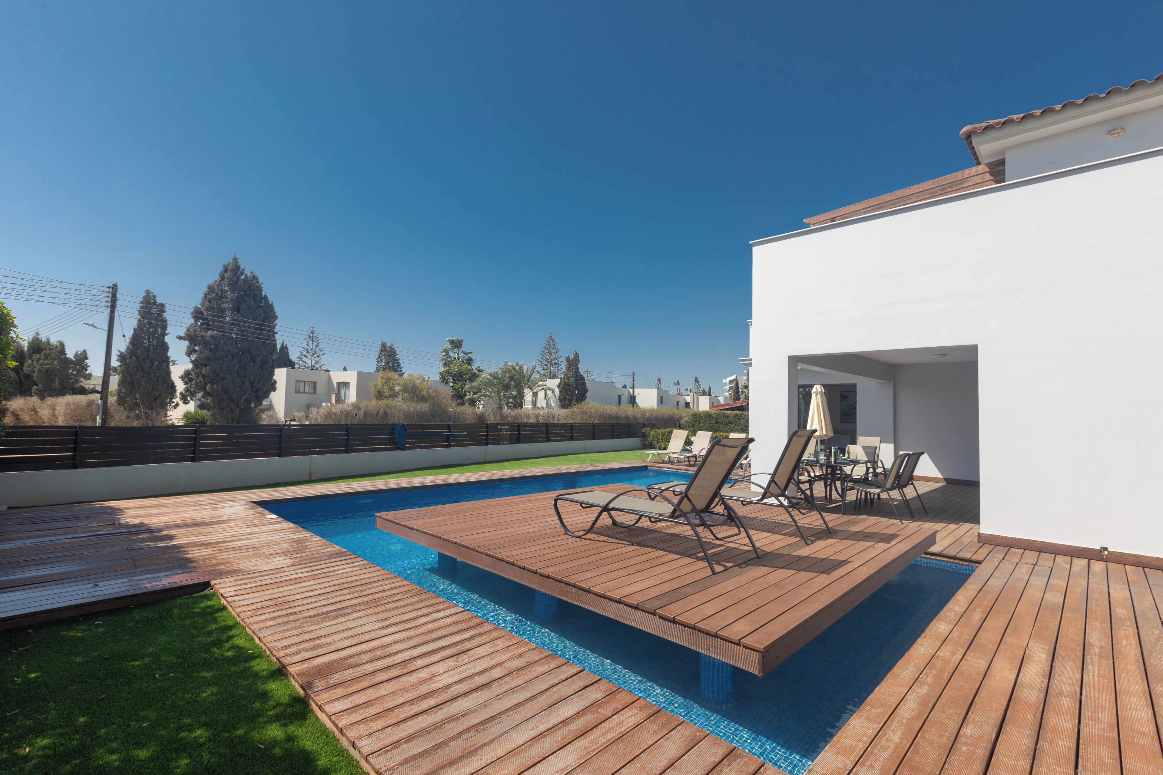 A modern outdoor pool area with wooden decking, lounge chairs, and a white building in the background.