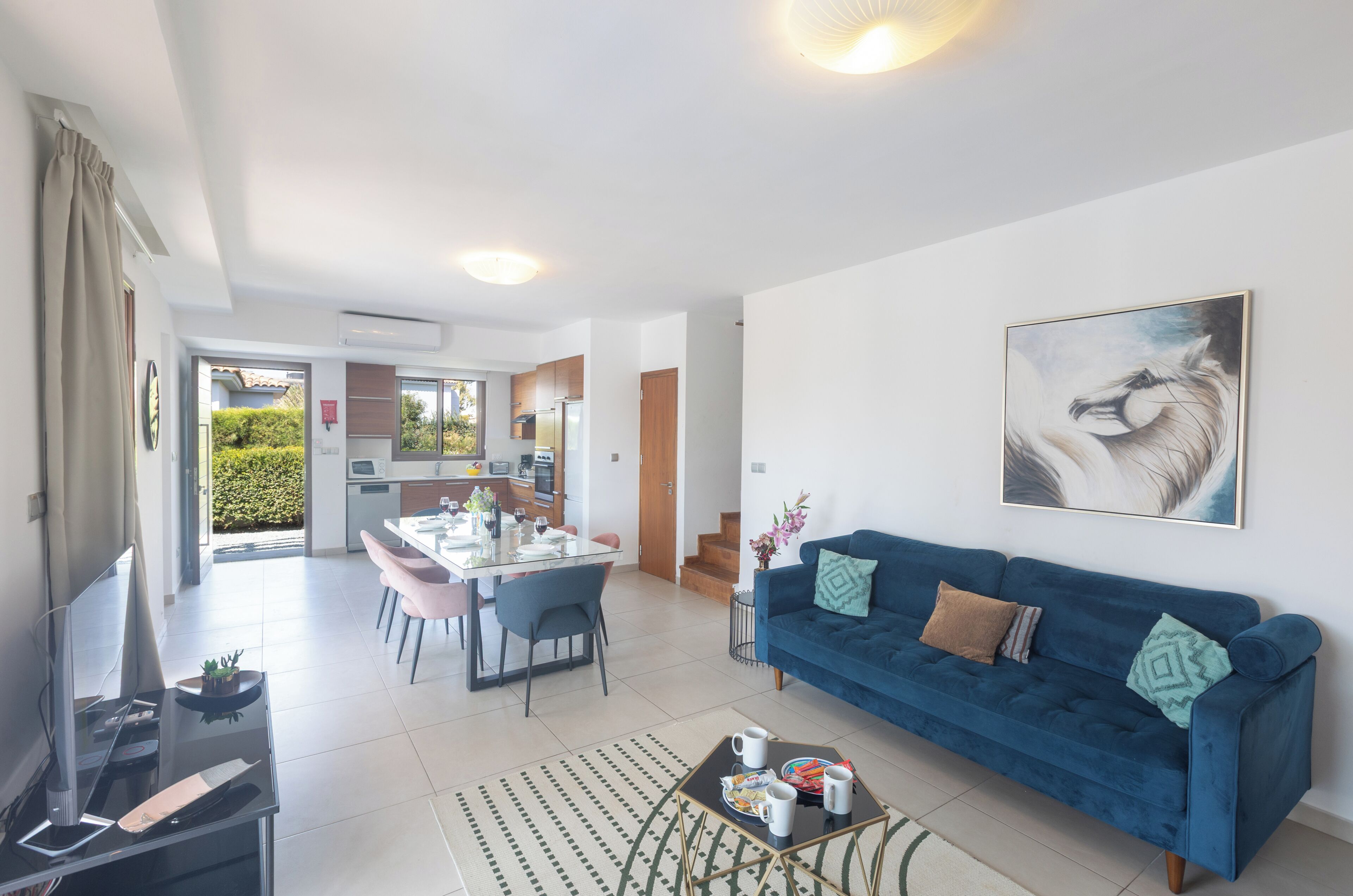 A modern living room with a blue sofa, a dining area with pink chairs, and a kitchen in the background.