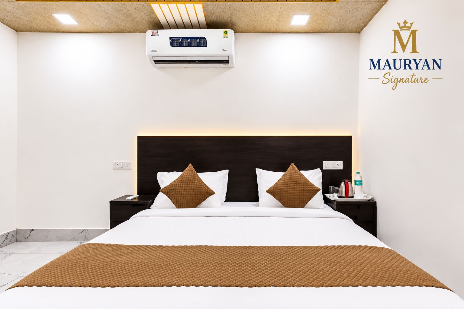 A hotel room with a bed, nightstands, an air conditioning unit, and a logo on the wall.