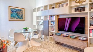 Una sala de estar moderna con una mesa de vidrio, sillas blancas, un televisor y una estantería llena de libros y objetos decorativos.