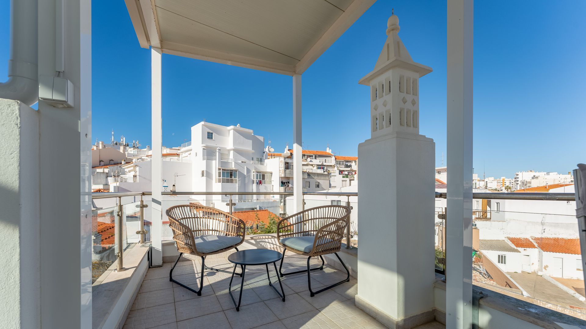A balcony with two wicker chairs and a small table, overlooking a cityscape with white buildings and a clear blue sky.