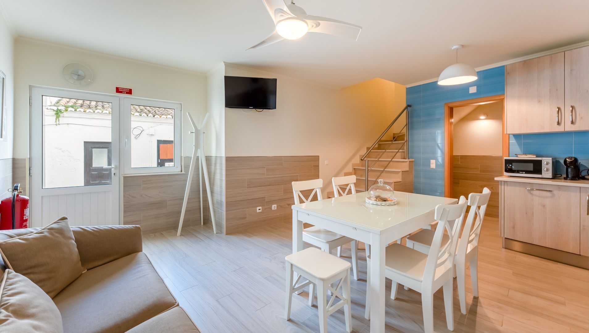 A modern kitchen with a microwave, toaster, and coffee maker. There is a dining area with a white table and chairs. A staircase leads to a second floor.