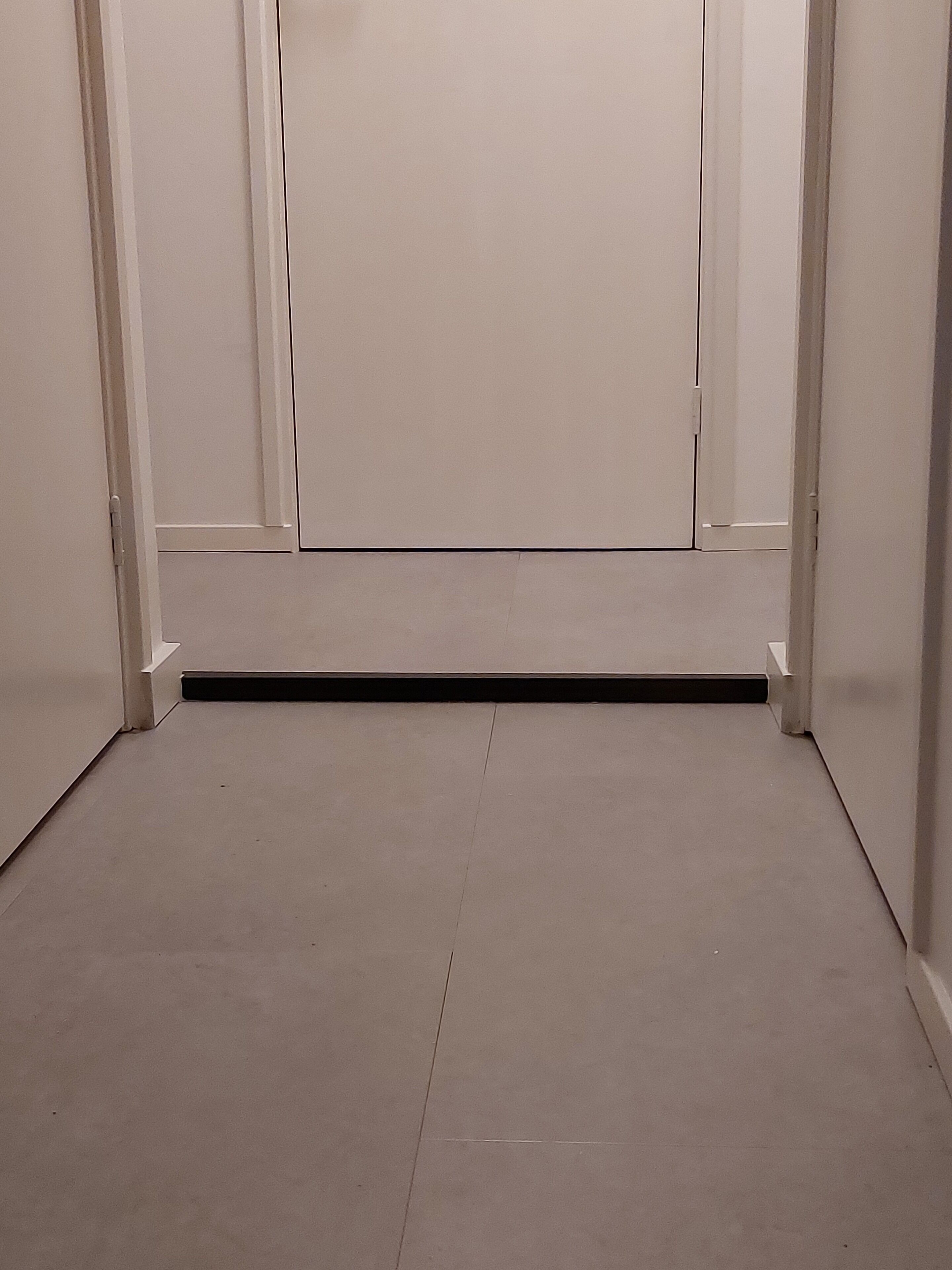 A room with a plain white door, a small vent on the floor, and white walls.