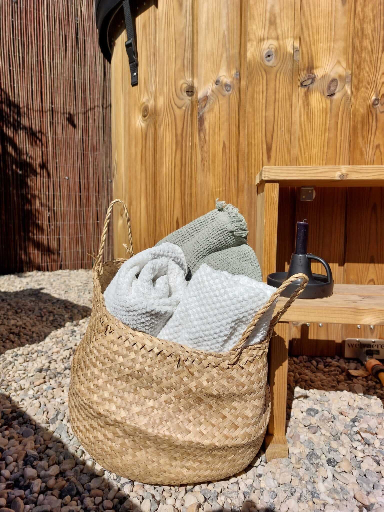Un panier en osier rempli de serviettes pliées, posé sur un sol de gravier devant une structure en bois, sur laquelle se trouve un objet noir, lui-même placé sur une étagère en bois.