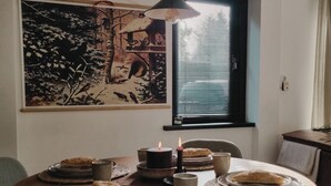Un coin repas avec une table ronde dressée pour deux personnes, agrémentée d’assiettes, de tasses et d’une bougie. Un tableau est accroché au mur, au-dessus de la fenêtre.