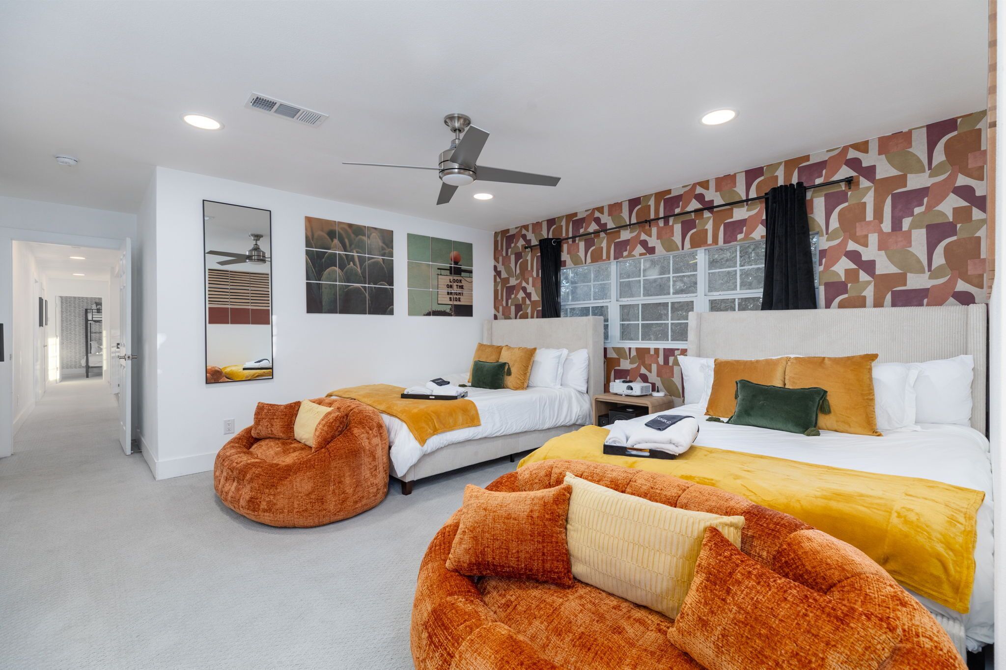 Quarto de hotel moderno com duas camas, ventilador de teto, parede estampada e uma janela grande.