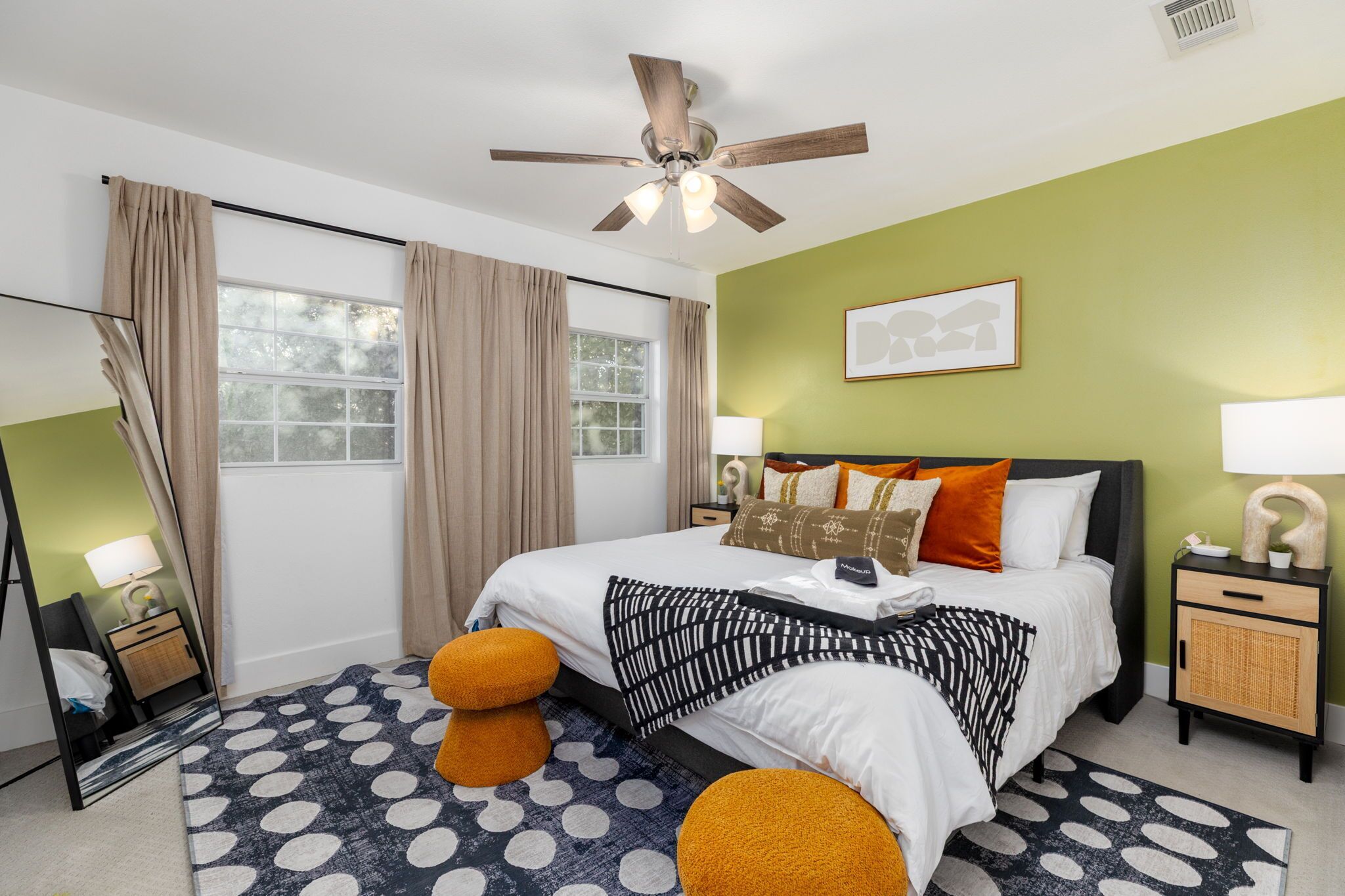 A modern bedroom with a large bed, a ceiling fan, a window with curtains, a nightstand with a lamp, and a mirror.