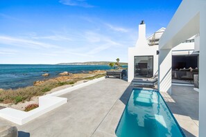 Ein moderner Poolbereich mit Blick auf den Ozean, ein Grillplatz und eine überdachte Lounge-Ecke.