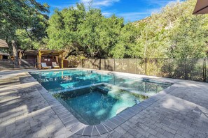 An outdoor swimming pool with a wooden deck and lounge chairs, surrounded by trees and a stone wall.