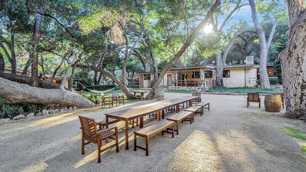 A rustic outdoor seating area with wooden benches and tables, surrounded by trees and a stone wall.