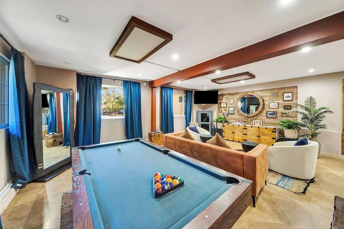Ein Billardtisch mit Spielzeug und Queue, eine Couch, ein Fernseher und ein Spiegel in einem Raum mit Holzbalken und einer Deckenleuchte.