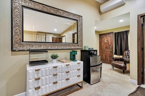 A modern living room with a large mirror, a white cabinet with drawers, a coffee machine, a refrigerator, a leather armchair, and a wooden door.