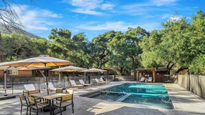 An outdoor pool area with lounge chairs and umbrellas, surrounded by trees and a wooden fence.
