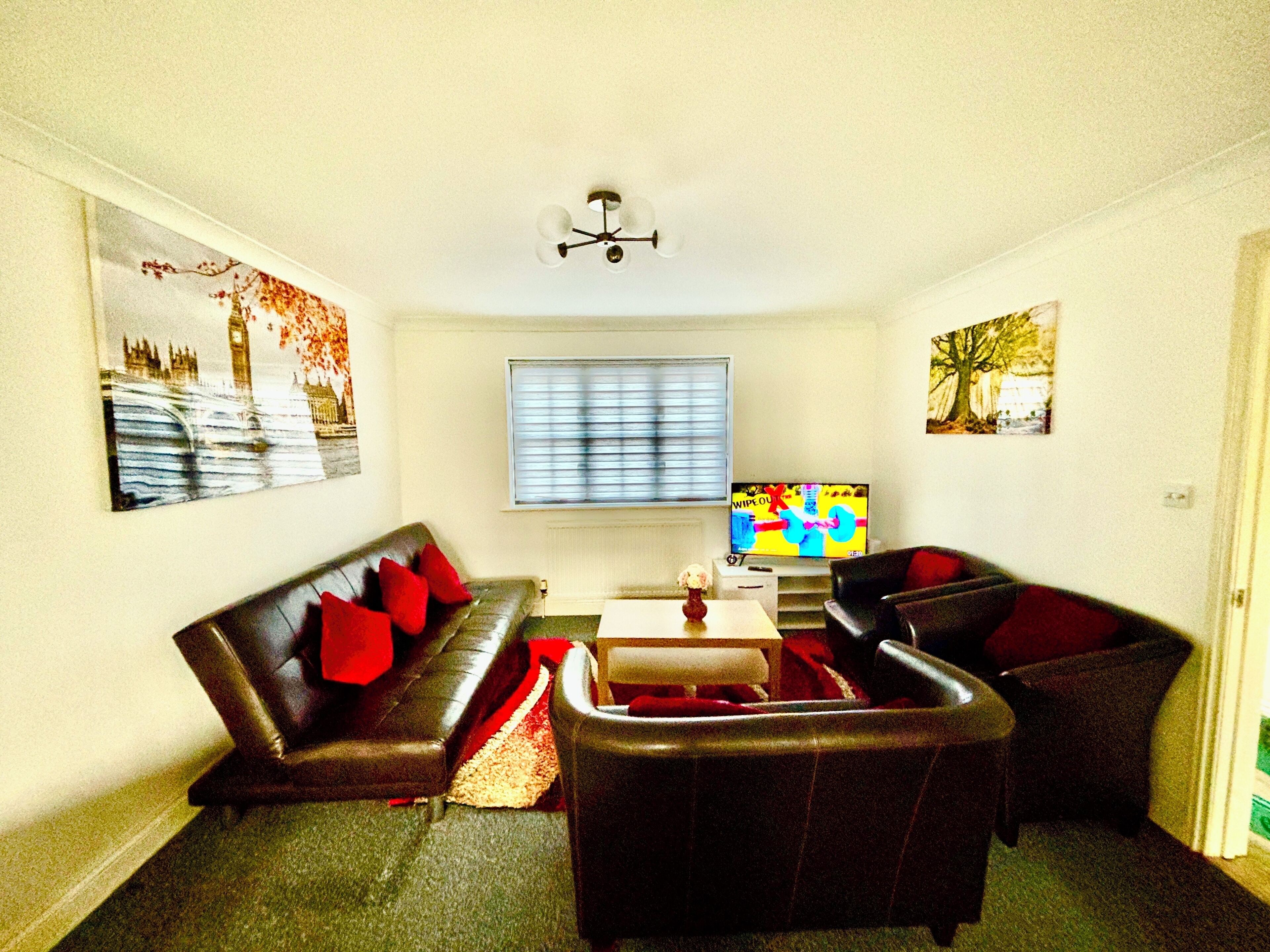 A living room with a sofa, armchair, and a coffee table. There is a window with blinds, a ceiling light fixture, and artwork on the walls.