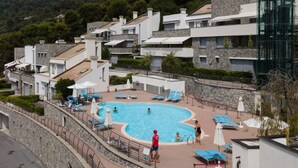 Ein Schwimmbeckenbereich mit Liegestühlen und Sonnenschirmen, umgeben von modernen Gebäuden mit Steinverkleidungen.