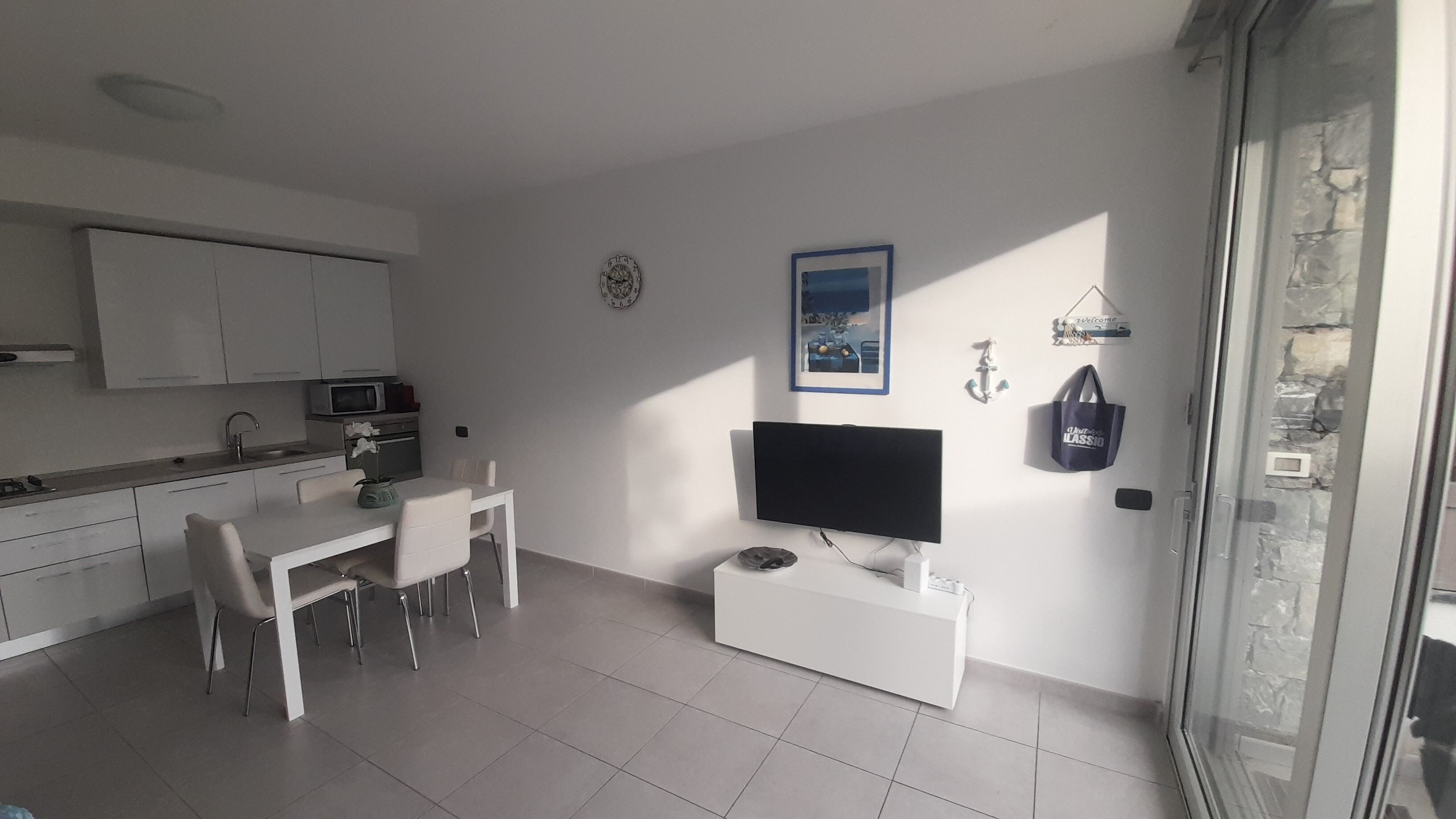 Eine moderne Küche mit weißen Schränken, ein Essbereich mit weißem Tisch und Stühlen, ein Flachbildfernseher auf einem weißen Schrank und eine Steinwand mit einem Fenster.
