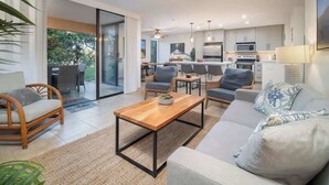 Una sala de estar moderna con sofá, mesa de centro y sillas, conectada a una cocina y comedor con un amplio ventanal corredero que da a un jardín.