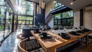 A modern dining area with a long wooden table set for a meal, surrounded by large windows offering a view of the outdoors.