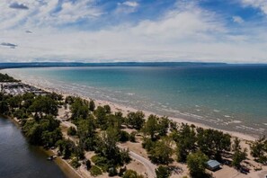 An aerial view of a beach with a road curving through a wooded area, leading to a sandy beach with clear blue water. - Full Cottage STEPS 2 BEACH Drive 2 Blue Mountain (Wasaga Beach)