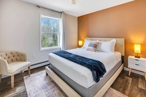 A bedroom with a large bed, a nightstand, a chair, and a window with a view of trees.
