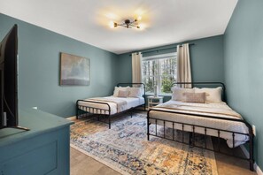 A bedroom with two single beds, a nightstand, a desk, a chair, a television, and a window with curtains.